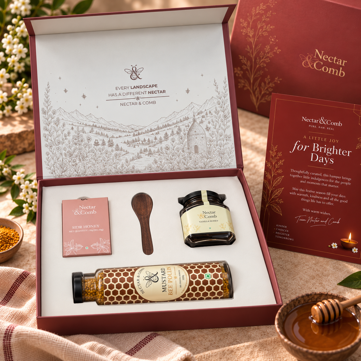 A luxury gift set from Every Landscape Honey & Lipped Nectar by Nectar & Comb, displayed in an open burgundy box. The set includes a honeycomb jar, a small jar of honeycomb spread, a pink box of after-sunset, a wooden spoon, and a booklet, all arranged on a rustic tablecloth with a honey pot and flowers in the background.