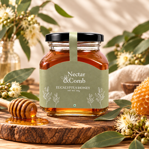A jar of Nectar & Comb Eucalyptus Honey sits on a rustic wooden slice surrounded by eucalyptus leaves, flowers, a honey dipper, and a honeycomb piece. The jar has a black lid and a sage green label with white text and botanical illustrations.