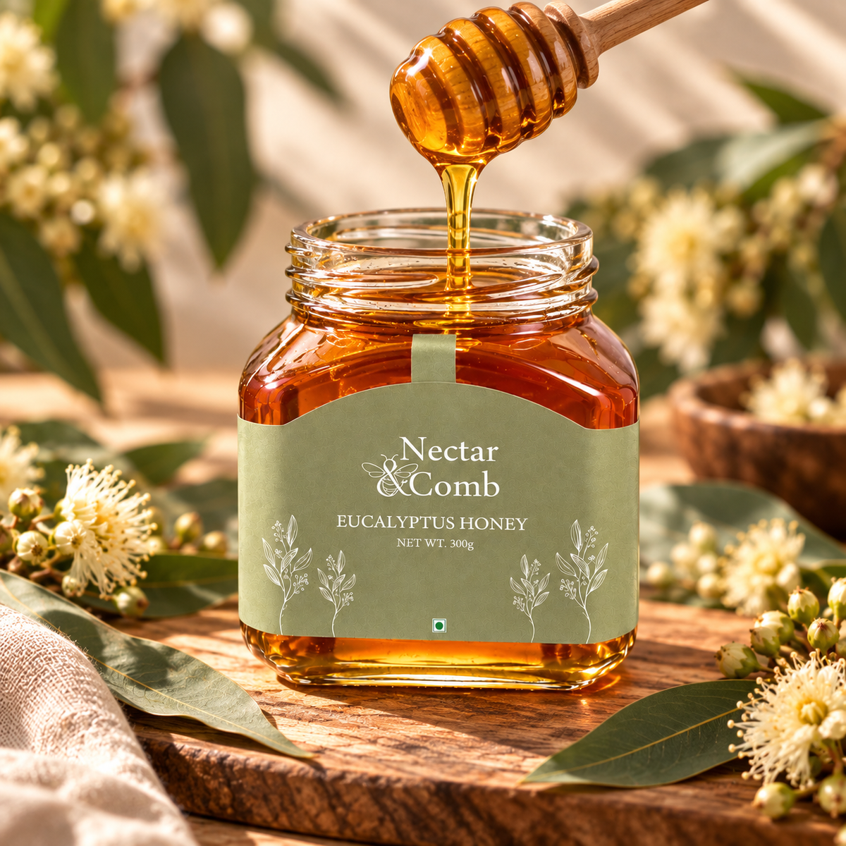 A glass jar of Nectar & Comb Eucalyptus Honey sits on a wooden surface surrounded by eucalyptus leaves and small white flowers, with honey dripping from a wooden honey dipper above the jar.