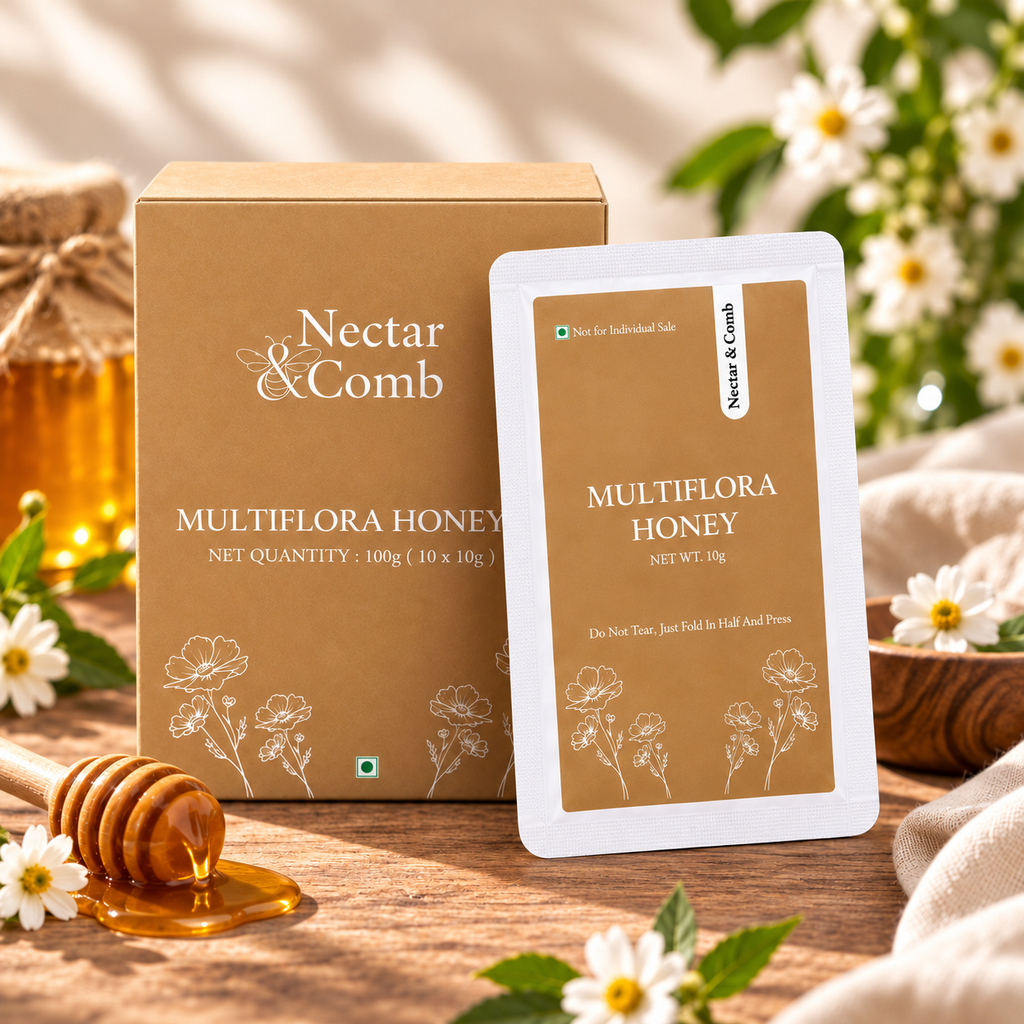 Nectar & Comb Multiflora Honey product shown with its brown box and a single honeycomb packet on a wooden surface, surrounded by white flowers, a honey dipper, and soft natural lighting.