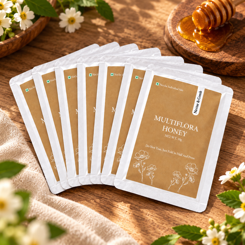 A fan arrangement of eight Multiflora Honey sachets on a rustic wooden table, surrounded by natural props like white flowers, a honey dipper, and a cloth.