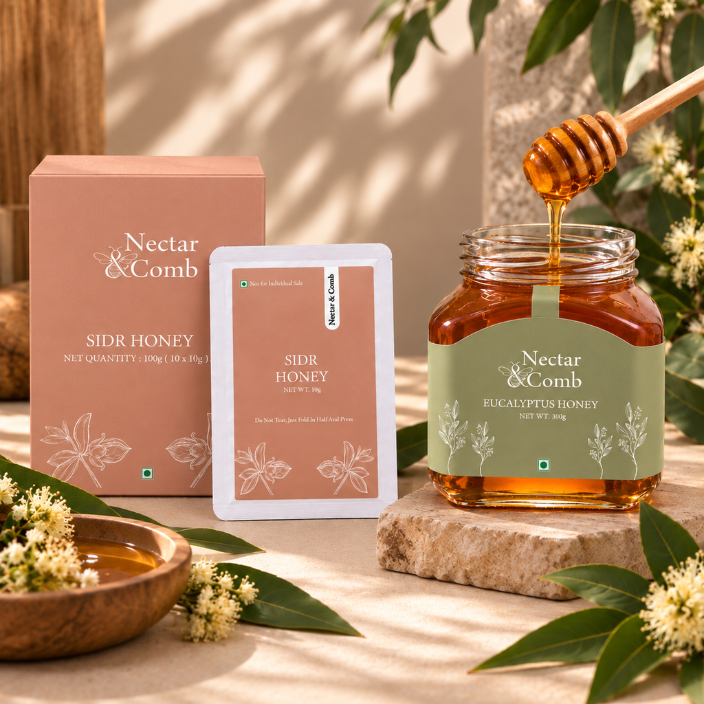 Nectar & Comb Sidr Honey eucalyptus honey jar with a wooden honey dipper, alongside its pink packaging box and a single packet, styled on a stone slab with eucalyptus leaves and a wooden bowl of honey.